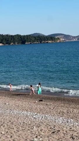 The video shows a sandy beach with several children playing near the water's edge. The water is blue and there are small waves coming to shore. In the background, there is a hilly landscape with trees. The sky is clear and blue, suggesting a sunny day. Some of the children are wearing swimsuits and light-colored clothing. The beach is made up of sand and small pebbles.