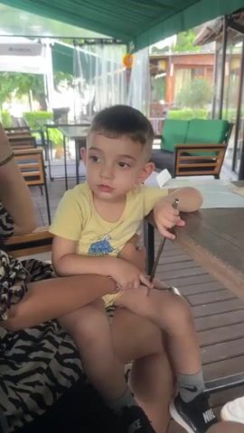 A young boy with short, dark hair sits on someone's lap at a wooden table outdoors. He wears a yellow t-shirt with a blue design, yellow shorts, grey socks with a black Nike logo, and black Nike sneakers. His left arm rests on the table, and he holds a fork in his right hand, which is near his mouth. He looks to his right and gestures with his left hand. The background shows a green awning, tables, chairs, and greenery, suggesting a restaurant or outdoor dining area. The camera is positioned at eye level with the boy.