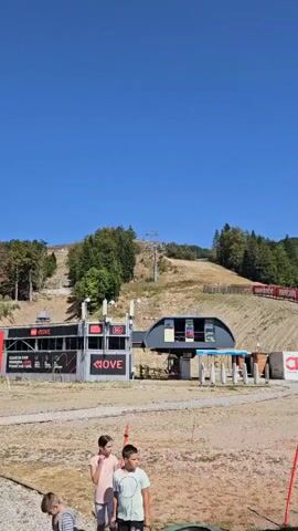 The video shows a cable car station and a ski slope on a sunny day. There are three children in the foreground, two girls and one boy, standing in a grassy area. Behind them is the cable car station, which has a sign that says "MOVE" and "5G". There are also several large antennas mounted on the station. Further up the hill is a ski lift with several cars. The ski slope is mostly bare, with some dry grass and scattered trees. The sky is clear and blue.