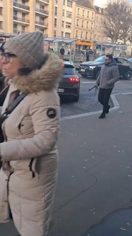 The video shows a street scene in a European city with light traffic. A man in a gray jacket walks past parked cars and a bicycle leaning against one of them. In the street, a gray car waits behind another, followed by two black Mercedes SUVs and a white car. Buildings line the street, and a bus stop is visible in the background. The sky is partially sunny. Car horns are heard.