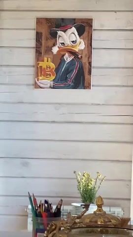 The video begins with a close-up shot of a painting of Scrooge McDuck wearing a hat and holding a Bitcoin symbol. The painting is hung on a wooden paneled wall. The camera then pans down to a desk with a container of colored pencils, a small plant, and a decorative gold object. The shot continues to pan, revealing a stack of folded towels, a clear bowl of water, and a blue box. Finally, the camera pans left to show an open doorway leading into another room, with sunlight streaming in. The overall environment appears to be a room with wooden walls and a desk. There are no people visible in the video.