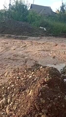This media file depicts a muddy, flowing body of water, likely a river or stream, in Uchbay, Kyrgyzstan. The scene is characterized by the movement of water over a bed of pebbles and dirt. In the foreground, there are piles of brown earth and stones, suggesting construction or natural erosion. In the background, there are trees and a building with a corrugated roof, indicating a rural or semi-rural setting. The lighting suggests it is either early morning or late afternoon, with a warm, golden hue illuminating the water and surrounding landscape. The overall impression is one of natural forces at play, perhaps a recent rainfall or a natural rise in the water level. There is no visible text in the media file.
