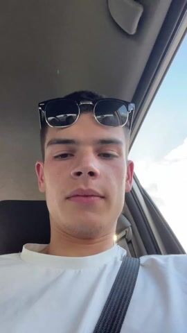 A young man with short dark hair and freckles sits in the front seat of a car, wearing a white t-shirt and a seatbelt. Sunglasses are perched on top of his head. The camera angle is slightly upward, suggesting he is holding a phone or camera above him. He looks at the camera and smiles slightly. He then turns the camera to the side to show a young woman sitting in the back seat. She has long, light brown hair, wears glasses, and is wearing a dark blue t-shirt with white trim. She appears to be pouting and waves briefly. The interior of the car is visible, with black seats and a light-colored ceiling. Outside the window, green foliage is visible, blurring as the car moves.