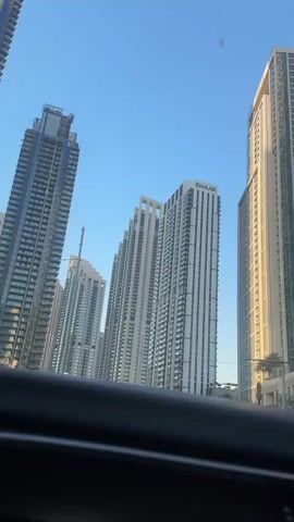 The video shows a view from inside a vehicle, looking out at a street lined with several tall, modern skyscrapers. The sky is clear and blue. The buildings are a mix of white and glass, with some appearing to be residential towers. One prominent building in the center has the Emaar logo visible. There are palm trees and some greenery along the sides of the street, suggesting an urban environment. The camera is moving forward, as indicated by the slight blur and the perspective shifting. There are no people or other vehicles clearly visible in the frame. The overall impression is of a bright, sunny day in a developed urban area.