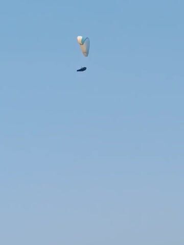 A paraglider is visible in the sky against a clear blue sky. The paraglider is white with blue accents and is carrying a person underneath. The paraglider appears to be descending. The camera then pans down to reveal ancient ruins overlooking the ocean. The ruins consist of stone walls and structures. The ocean is a vibrant blue and is calm. The sky is clear and hazy in the distance.