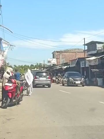A motorcycle is parked on the left side of the road. A person in a white head covering walks away from the motorcycle, followed by another person also in a white head covering. A dark colored car is driving down the road, and behind it, a motorcycle is pulling a cart with cargo. A black car is driving towards the viewer on the right side of the road. Several buildings with open fronts line the right side of the street. The sky is clear and blue.