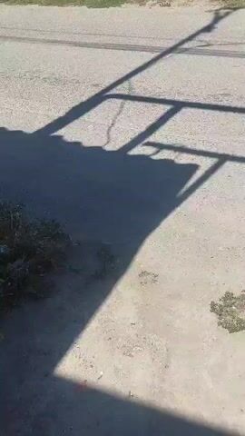 The video shows a street with buildings on either side. The street appears to be paved and there are tire marks visible on the surface. There are also shadows cast on the road, indicating it is daytime. Some of the buildings have walls made of stone or concrete, and some have roofs made of corrugated metal or tiles. There are power lines and street lights visible along the street. In the distance, a small truck or van can be seen. There is some vegetation, such as grass and bushes, growing along the sides of the road and in front of the buildings.