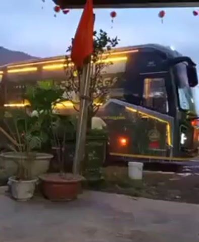 The video shows two buses facing each other. The bus on the left is dark green with illuminated trim, while the bus on the right is yellow with red trim. Both buses have their headlights on. Two men stand between the buses, near the right bus. One man wears white pants and a light-colored shirt, while the other wears darker clothing. Water streams between the two buses. The environment includes plants in pots, a small white bucket, and red lanterns hanging overhead. The scene appears to be outdoors, with a glimpse of the sky in the background.