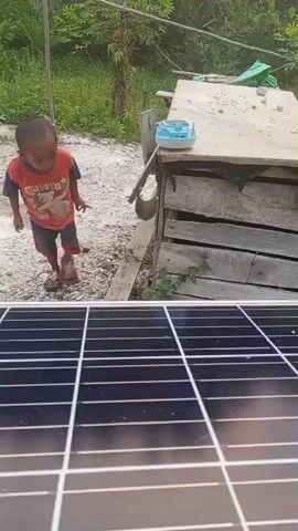 A child stands in front of a small wooden structure. The child is wearing an orange and blue shirt and black shorts. They are looking down at something. In the foreground, a solar panel is visible. The background consists of greenery and a dirt path. A small blue object is on top of the wooden structure.
