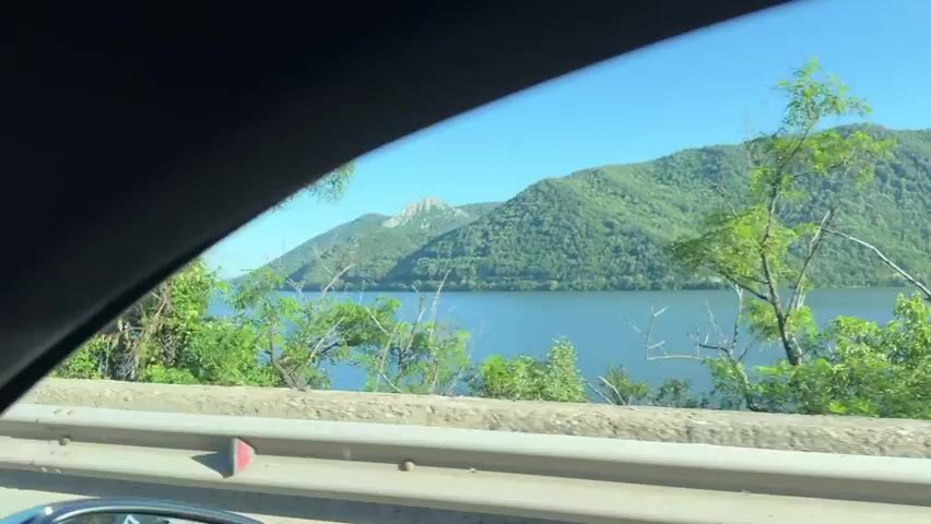 The video shows a view from inside a moving vehicle, looking out the side window. A bright blue sky with no clouds is visible at the top of the frame. Below the sky, there is a wide body of water, likely a lake or river, which is also a bright blue color. On the far side of the water, a range of green, tree-covered mountains stretches across the horizon. In the foreground, a gray concrete barrier or guardrail runs horizontally across the bottom of the frame. Beyond the barrier, there are various green bushes and trees, some appearing close to the vehicle and some further away, partially obscuring the view of the water and mountains. The vehicle is moving, indicated by the continuous change in the landscape as it passes by. The overall scene depicts a scenic natural environment on a clear, sunny day.