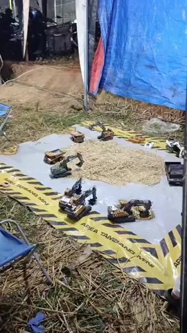 The video shows a collection of toy construction vehicles arranged on a tarp, which is laid out on the ground. The tarp is marked with yellow and black hazard tape that reads "AREA TAMBANG MINI". A pile of light-colored granular material, possibly sand or gravel, is in the center of the tarp, surrounded by the toy vehicles. Several toy excavators and dump trucks are visible. The background is dimly lit and appears to be an outdoor setting, possibly a yard or an open area. A blue tarp is visible in the background, partially covering something. There are also some dry stalks of hay or straw on the ground around the tarp. A blue chair is visible in the lower left corner. There are a few people partially visible in the scene, including someone in the lower right corner wearing a white shirt. The overall impression is a miniature construction site setup for play or display.