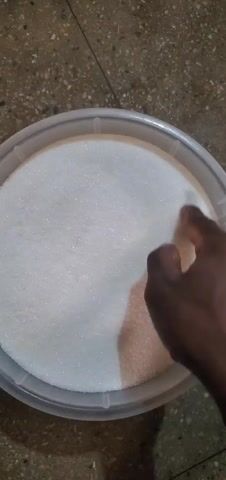A hand is moving in a circular motion inside a round, plastic container filled with granulated white sugar. The hand has dark skin. The container is light-colored. The background is a speckled, light brown surface.
