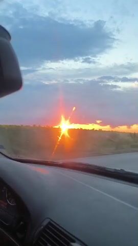 The video shows a view from the interior of a car driving on a road. The sun is setting in the distance, casting a warm orange and pink glow across the sky. Clouds are scattered across the sky, some illuminated by the setting sun. The landscape outside the car consists of fields and trees. The road is paved and appears to be in good condition, with white lines marking the lanes. The car is moving at a moderate speed. The sun's rays are bright and create lens flares on the windshield. There are no people visible in the video.
