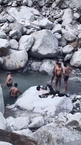 The video shows a group of men bathing in a rocky river. Clothes are laid out on a large rock near the water. The surrounding environment is mountainous with lush green hills and a clear blue sky. The men appear to be enjoying themselves in the river.