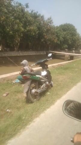 A video shows a person riding on a motorcycle. The view is from the perspective of the rider, looking forward and slightly to the right. The rider's hand is visible on the handlebar, and a side mirror is also in view, reflecting the rider and the surrounding environment. The motorcycle appears to be moving along a paved road that runs parallel to a canal or a ditch filled with murky water. On the left side of the canal, there are trees and some buildings, suggesting a rural or semi-urban area. The ground alongside the road is covered with grass. The sky is visible in the background, indicating it is daytime. The video is shaky, suggesting the motorcycle is in motion.