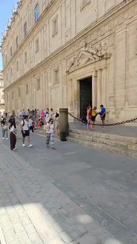 The video begins with a view of a large, light-colored stone building with a grand entrance. Several people are visible near the entrance, some standing and talking, others walking. The camera then pans to the left, showing a street with a sidewalk and tram tracks. More people are walking on the sidewalk, and a man is riding a bicycle in the distance. The video continues to show a street scene with buildings on either side, people walking, and tram tracks. The day appears to be sunny.