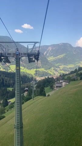This media file captures a scenic view from a cable car ascending a lush green mountainside. The cable car itself is a prominent object, with its supporting tower and cables dominating the foreground. Several other cable car cabins are visible suspended along the cable, indicating that this is an operational ski lift or gondola system, likely used for transportation up the mountain.

The scene is set in a mountainous region, with rolling green hills covered in grass and scattered with trees. In the distance, a valley unfolds, revealing a charming village with houses and buildings clustered together. The background is filled with majestic mountains, their slopes a mix of green forests and rocky outcrops. The sky is a clear blue with a few wispy white clouds, suggesting a pleasant day.

The time of day appears to be daytime, likely late morning or early afternoon, given the bright sunlight casting shadows. The weather seems to be warm and sunny, perfect for outdoor activities. The location context provided places this scene in Urtijëi - St. Ulrich in Gröden - Ortisei, Italy, which is known for its stunning alpine scenery and ski resorts.

While there are no clearly identifiable people in the foreground, the presence of the cable car cabins suggests that people are being transported or have been transported. The overall impression is one of natural beauty, peacefulness, and recreational activity. The vibrant green of the landscape and the clear blue sky evoke a sense of awe and tranquility. No visible text is present in the media file.
