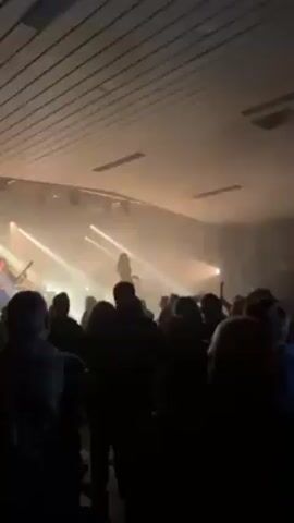 The video shows a live band performing on a brightly lit stage in front of a large audience. The stage is filled with fog, obscuring some of the performers, but it’s possible to make out a vocalist and a guitarist. The audience is standing and appears to be enjoying the performance, with some individuals raising their hands and moving to the music. The venue appears to be a large hall with a simple, utilitarian ceiling. The overall atmosphere suggests an energetic and lively concert.