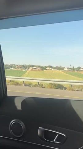 From the perspective of a passenger in a moving vehicle, the video depicts a scenic view through the car window. Outside, a rural landscape unfolds, featuring a green field, possibly a farm or agricultural area, and houses in the distance. A gray guardrail and a highway with a yellow line separate the road from the field. A green road sign with white arrows is visible, suggesting directions or lane information.  The sky is clear and blue. The interior of the car is visible, including the window frame, door panel, and a speaker.