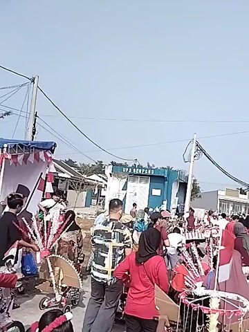 The video shows an outdoor scene with a clear sky and some overhead wires. A group of people are gathered on a paved area, possibly a street or courtyard. Many individuals are wearing red and white clothing, and red and white flags are visible, suggesting a patriotic or celebratory event. Several people are pushing or riding on decorated bicycles, some adorned with flags and festive items. In the background, there are buildings, including one with a sign that reads "UP3 BEKASI." The atmosphere appears to be lively and communal, with people interacting and participating in what seems to be a local parade or festival.