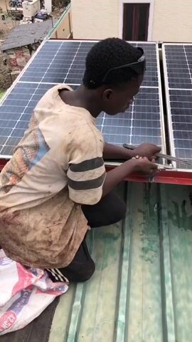 A young man is installing solar panels on a rooftop. He is wearing a dirty, light-colored t-shirt with black stripes on the sleeves and black pants. He has sunglasses on top of his head. He is kneeling on the roof, which is made of corrugated metal. In front of him is a white bag with red and blue markings, and what appears to be a bag of cement. He is holding a tool, possibly a wrench or screwdriver, and is working on the frame of a solar panel. The solar panels are mounted on a red metal frame. Another person, a man in a blue shirt and dark pants, is visible in the background, also working on the solar panels. There are some wires connected to the panels. The background shows a building and some trees.