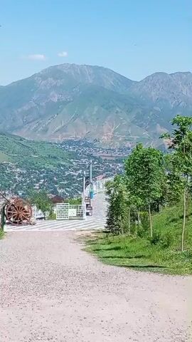 The video shows a mountain scene on a sunny day. Initially, a dirt path is in the foreground, leading to a paved area with a white fence. In the background, there's a panoramic view of a town nestled in a valley, framed by large mountains. The camera then pans slightly to reveal a large, white, arched structure surrounded by green grass and small trees. Following this, the camera returns to the initial viewpoint, where three people are walking down the path towards the paved area. The mountains and the town remain visible in the background.