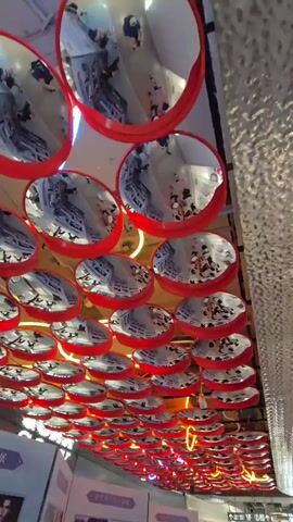 A low-angle shot captures a ceiling installation of numerous circular mirrors, each with a red frame, suspended in rows. The mirrors reflect an interior space, likely a store, showing people walking and browsing among displays. The overall impression is one of a busy and visually dynamic environment, with the mirrors creating a sense of expanded space and fragmented imagery. Neon lighting in a yellow-orange hue is visible along the edges of some of the lower mirrors, adding a modern and vibrant touch to the scene. The ceiling itself appears to be dark, with exposed beams and ventilation elements.