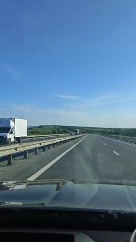 The video shows a car driving on a highway on a sunny day. The sky is blue with a few scattered clouds. The highway has multiple lanes and a guardrail on the side. A large truck passes the car, and there is a highway sign visible in the distance. The landscape surrounding the highway includes green fields and trees. The video is taken from the perspective of the driver, showing a portion of the car's dashboard.