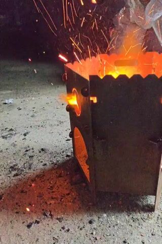 This media file depicts a metal portable grill or brazier, actively in use, with flames and sparks emanating from its top. The grill is dark, likely made of steel, and features a decorative, scalloped top edge. Circular openings are visible on the sides, through which the intense heat and light of the fire are visible. The grill rests on a surface that appears to be dirt or gravel, with scattered embers and ash around its base, indicating it has been used for a fire. In the background, the scene is dark, suggesting it is either nighttime or a dimly lit area. There are some indistinct shapes in the background, possibly bags or other objects. The ground surface is a mix of gravel and dirt with scattered small red embers. The overall impression is one of an outdoor setting where a fire is being maintained for cooking or warmth. The sparks suggest an active and perhaps breezy environment. The location context of Svetly, Russia, places this scene in a specific geographic and climatic setting, though no direct visual cues confirm this without the provided context. No text is visible in the media file.
