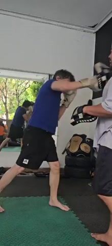 Two men are practicing Muay Thai in a gym in Bekasi, Indonesia. One man, wearing a blue t-shirt and black shorts, is throwing punches while wearing white boxing gloves. The other man, wearing a gray t-shirt and black shorts, is holding black and white padded mitts to block the punches. There is a large black banner on the wall with red lettering that reads "MUAY THAI EST. 2021 R..." The gym floor is covered with green and black mats. In the background, a mirror reflects other people training, one of whom is wearing an orange shirt. Various boxing gloves and protective gear are visible on the floor and on shelves. The lighting suggests it is daytime.