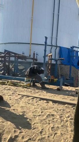 A person wearing a dark jacket, grey pants, a reflective vest, and a welding helmet is welding a metal pipe. The person is seated on a yellow bucket. They are working outdoors on a sandy surface. In the background, there is a large white cylindrical structure and some blue tarps. There are also metal structures and pipes in the mid-ground. The sun is casting long shadows, indicating it is likely morning or late afternoon.