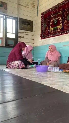 A group of women and one child are sitting on the floor in a room. The room appears to be a simple dwelling with wooden walls and a tiled floor. There are tapestries hanging on the walls, and a window is visible. The people are dressed in casual clothing, with some of the women wearing headscarves. They appear to be engaged in a conversation, with some holding papers and one person speaking. There are also some dishes and glasses on the floor.