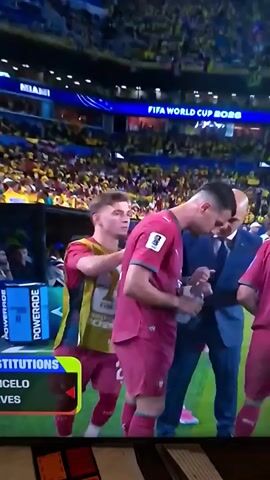 A soccer player in a maroon uniform is being spoken to by a man in a suit. Another player in a maroon uniform with the number 13 on his back stands to the right. A player in a yellow and maroon vest stands to the left, with his hand on the player in the maroon uniform. The players are on a soccer field with spectators in the background. A "FIFA WORLD CUP 2026" sign is visible.