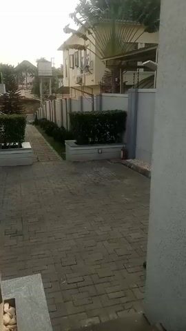 The video shows a view from a slightly elevated position, likely a balcony, looking down into a courtyard or driveway area. The courtyard is paved with gray bricks in a geometric pattern. On either side of the courtyard, there are hedges planted in raised concrete planters. The hedges are dense and green. Beyond the hedges, there are tall white buildings with black windows. In the distance, one can see a water tower and more greenery. The camera moves slightly during the video, changing the viewing angle. The lighting suggests it is daytime, possibly early morning or late afternoon.  A metal railing is visible in the foreground.