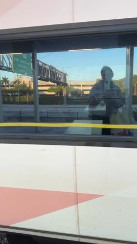 The video opens with a shot from inside a train, looking out a window. The reflection of a person wearing a dark top and carrying a bag is visible in the glass, holding what appears to be a phone. Outside, a sunny, clear sky is visible, along with a large bridge structure (possibly a railway or elevated road bridge), trees, and distant buildings. Mirrored text like "220" and "yewes nemes m" is visible, likely from an overhead road sign. A yellow stripe runs horizontally across the bottom of the window frame. The interior wall of the train features a geometric pattern of white, pink, and dark shapes.

As the camera pans slightly, the train doors begin to open. The exterior of the train is revealed to be white with black and pink/orange stripes, bearing the word "brightline" in black font and a large number "4" with a yellow accent. A yellow, textured train platform is visible below.

The doors fully open, and a person in a dark blue hoodie and dark pants, matching the reflection seen earlier, exits the train. They are wearing a black backpack and pulling a small, black, wheeled suitcase. Inside the open doorway, an illuminated ramp extends from the train to the platform. The interior of the train is bright, and other passengers can be seen seated inside. The side of the train also shows the numbers "3" and "215". The person steps onto the yellow platform, completing their disembarkation. The scene is well-lit, suggesting late afternoon or early evening.
