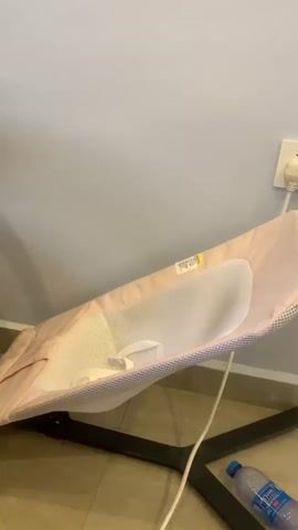 The video shows a pink baby bouncer leaning against a light blue wall. A black office chair is to the left of the bouncer. A white cord runs from the bouncer to an electrical outlet on the wall. A plastic bottle of water is on the floor next to the bouncer. The bouncer appears to be empty. The lighting is dim.