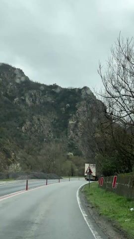 The camera, inside a vehicle, records a winding paved road on an overcast day. The road is marked with a white line on the right side and a red line on the left. A white semi-trailer truck moves ahead in the same direction. Road signs are visible on the right, including one warning about falling rocks and another restricting vehicles from passing, indicating a distance of 14,300 meters. On the left, a mountain covered in gray rock and sparse vegetation rises up. Bare trees stand along the roadside. The sky is uniformly gray, and the ambient noise includes music.