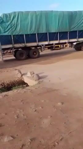 A large truck covered with a green tarp is parked on a dirt road. A motorcycle passes by, and then a car pulls a motorcycle behind it. Another motorcycle with three people on it drives by. The scene is a dusty, outdoor area with a clear sky.