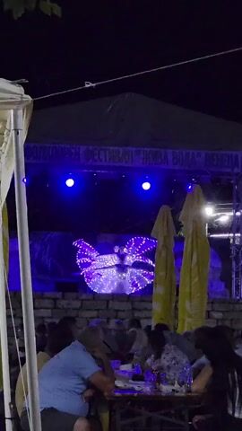A performance is taking place on a stage at night. The stage is lit with bright blue lights, and a performer in a sparkling, elaborate costume is dancing. The performer's costume has large, wing-like structures that are adorned with many small lights. Behind the performer, there appears to be a backdrop with a naturalistic scene, possibly a landscape.

In the foreground, an audience is seated at tables, seemingly at an outdoor venue. People are eating and watching the performance. Several large, yellow umbrellas are visible, providing shade or cover for the audience area. The overall atmosphere appears to be festive and engaging.

Above the stage, a banner with text is visible. The text is in Cyrillic script and reads: "ФОЛКЛОРЕН ФЕСТИВАЛ \"НИВА ВОДА\" ВЕНЕЦИА" which translates to "Folklore Festival 'Niva Voda' Venezia".

The time of day is clearly night, indicated by the darkness of the sky and the artificial lighting of the stage and venue. The weather seems to be pleasant, as people are gathered outdoors. The location is suggested by the banner text referring to a festival. The emotions conveyed are likely enjoyment and interest from the audience.