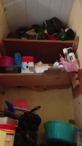 The video shows the inside of a cluttered, wooden storage space with three shelves. The top shelf holds dark-colored bags. The middle shelf contains various items, including a red and white plastic container, a blue cloth, and assorted small containers. The bottom shelf has more diverse items, such as a yellow and red container, cookware, a black plastic bag, and a green plastic basin. The storage space appears to be in a somewhat dilapidated state.