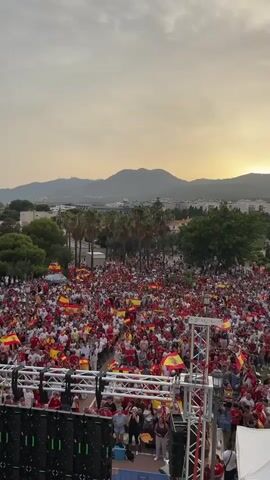 The video shows a large crowd of people gathered outdoors, many waving Spanish flags. The setting appears to be a town or city with buildings and palm trees visible in the background, and mountains in the distance. The sky is overcast with a warm, sunset glow. In the foreground, there is a stage or platform with equipment, including large speakers. People are standing and milling around, and some are holding up their phones, suggesting they are filming or taking pictures. The overall atmosphere seems to be one of celebration or a public event.