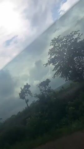 The video shows a view from a moving vehicle, likely a car. The camera is angled upwards, capturing a landscape dominated by green foliage and a cloudy sky with visible sun rays. There are several trees, some silhouetted against the bright sky, and a hillside covered in vegetation. The ground in the foreground appears to be a dirt or gravel road. The overall impression is of a natural, rural environment, possibly during a partly sunny or overcast day.