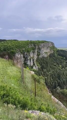 The video shows a view from a cliff edge. A wire fence runs along the edge, with green grass and wildflowers in the foreground. Across the fence, there's a steep drop to a forest of green trees. A large, flat-topped cliff face is visible, covered in vegetation on top. In the distance, there are green fields and a glimpse of what appears to be the horizon under a cloudy sky. The overall scene depicts a natural landscape with varied topography.