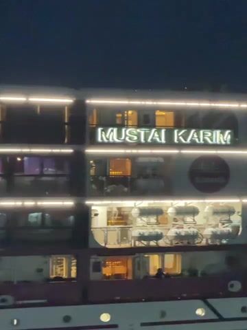 A video captures a large, multi-decked vessel at night. The vessel is illuminated with bright white lights along its edges and warm interior lights emanate from its windows. Visible on the side of the vessel is prominent signage that reads "MUSTAI KARIM." Life rafts are visible on one of the decks. The vessel is situated on dark water, likely a river or harbor, with reflections of the lights shimmering on the surface. The overall scene suggests a passenger ship or cruise liner at night.