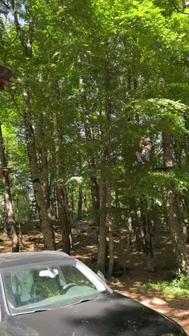 A medium shot in a forested adventure park shows a young girl on a zip line. She wears a white helmet, pink top, and black-and-white checkered pants, holding onto the line with both hands, facing left as she glides from right to left. Below her, a black car is parked facing left, partially visible. Behind her, a white car is parked, facing right. Tall trees with lush green leaves fill the background, with platforms and other zip lines visible among them. Sunlight filters through the canopy.