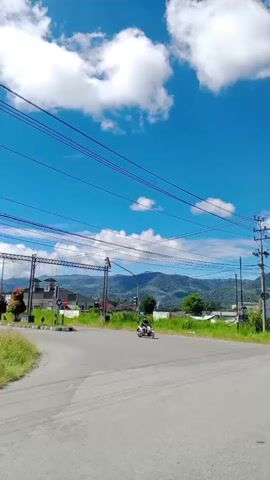 A motorbike is seen from the side, with a person on it, riding through a road intersection. The sky is blue with white clouds, and there are mountains in the background. There are power lines running across the sky. A white SUV is seen driving on the road ahead, and then multiple motorbikes are seen in a line on the road. The weather appears to be sunny and clear.