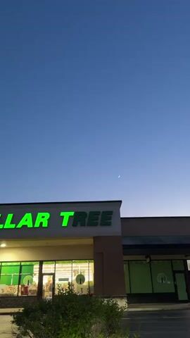 The video begins with a view of the exterior of a Dollar Tree store at dusk. The store sign is illuminated with bright green lettering. Inside the store windows, merchandise is visible, including circular displays with what appear to be clocks. To the right of the main store entrance, there is another storefront with green paneling and a Dollar Tree logo. Above the store, the sky is a deep blue, gradually lightening towards the horizon. A crescent moon is clearly visible in the sky, accompanied by a small, bright point of light, likely a planet such as Venus. Some green foliage is in the foreground, obscuring the bottom portion of the building. The camera pans slightly upwards, focusing more on the sky and the celestial bodies. The video ends with a close-up of the moon and the accompanying planet against the twilight sky, with the top of the Dollar Tree building still in view at the bottom of the frame.