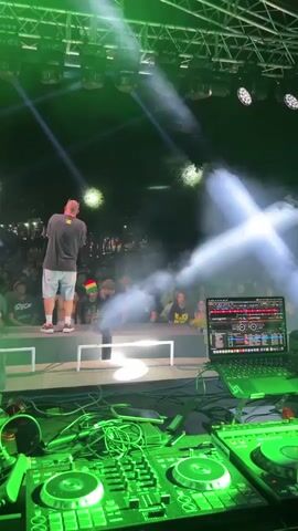 The video shows a stage at a night concert. The stage is lit with bright green lights. On the stage, a man is standing on a raised platform, facing away from the camera, and appears to be speaking into a microphone. He is wearing a dark t-shirt and light-colored shorts. In front of the stage, a DJ setup is visible, including a laptop with DJ software open and two turntables. A crowd of people can be seen in the background, some holding up their hands and illuminated by the stage lights. There is smoke or fog effect on the stage, catching the light and creating beams.