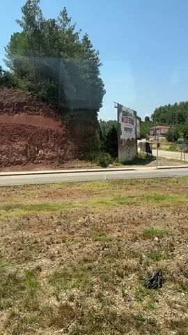 This media file captures a drive through a seemingly rural or suburban area in Osmaniye, Turkey, on a clear, sunny day. The scene unfolds along a paved road bordered by dry, grassy terrain and a steep, reddish-brown embankment on the left, covered in sparse vegetation. The background is dominated by lush green trees, suggesting a hilly or mountainous landscape.

Several key objects and visual cues indicate an urban or semi-urban setting. A large billboard stands prominently on the left, displaying text and images, although the details are difficult to discern. Further along, a traffic intersection is visible, complete with traffic lights, pedestrian crossing signs, and decorative landscaping featuring small, conical trees.

A significant focal point is a large, decorative sign in the middle of a roundabout. The sign spells out "OSMANİYE" in golden, three-dimensional letters, with a red heart and a cartoonish yellow figure integrated into the design. This "I love Osmaniye" style monument is a clear location marker. Smaller signs, including one with the word "VEKİL" and another with what appears to be a political advertisement, are also visible. Buildings in the distance suggest residential or commercial establishments.

The time of day is clearly daytime, indicated by the bright sunlight and long shadows. The weather appears to be warm and fair, with a clear blue sky. The overall impression is one of a transition from a natural landscape to a developed area, likely a town or city entrance, marked by civic pride and infrastructure. No people are clearly visible, but the presence of traffic signals and pedestrian signs implies human activity and movement.