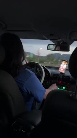 The video shows the interior of a car at dusk. The camera is positioned in the back seat, facing forward. A person with long dark hair is driving, wearing a blue t-shirt and jeans. Their left hand is on the steering wheel, and their right hand is near the car's radio. A smartphone is mounted on the dashboard, displaying a grid of icons. Outside the car, trees and signs are visible, suggesting a highway or road setting. The car's dashboard lights are on, and the radio display is illuminated in green. The driver's reflection can be seen in the rearview mirror.